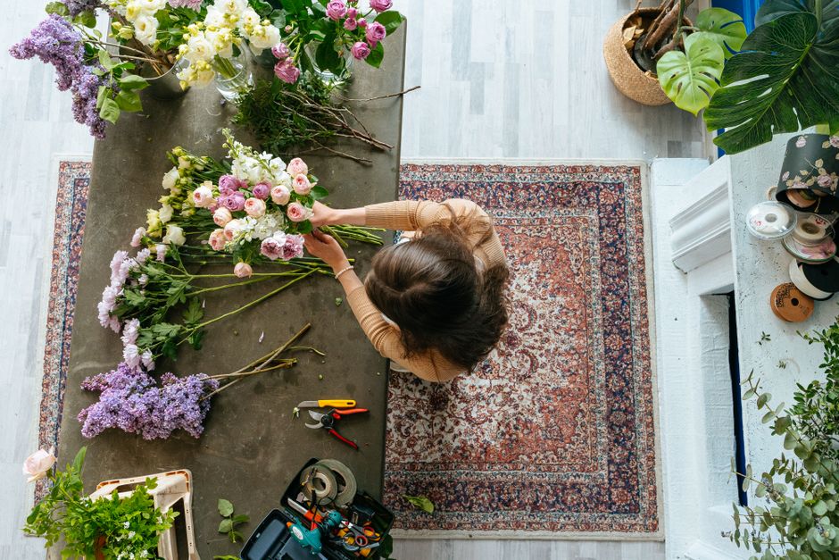 A top florist on Fulham Road arranging a bouquet of fresh flowers on a grey work table in a well-lit, cozy floral studio. The bouquet features pink and white roses, complemented by purple lilacs and w