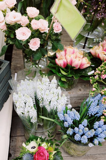 A display of fresh floral arrangements featuring pale pink roses in a clear glass vase, alongside pink and yellow tulips in a round bouquet with green foliage. Surrounding these, there are blue and wh