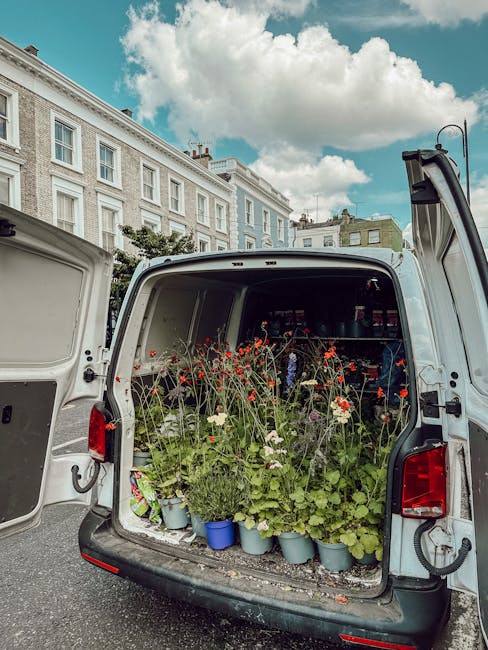 The open rear of a white van filled with flowering plants and potted flowers, including red poppies and white daisies, arranged in blue and green pots. The van is parked on a street with a backdrop of