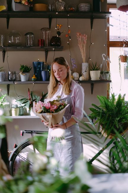 A young woman with long, light brown hair, wearing a white shirt and apron, is standing indoors in a well-lit room with wooden shelving decorated with potted plants, vases, and floral arrangements. Sh